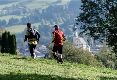 Two runners are jogging across a green meadow. In the background, gentle hills and a small village with a church can be seen.