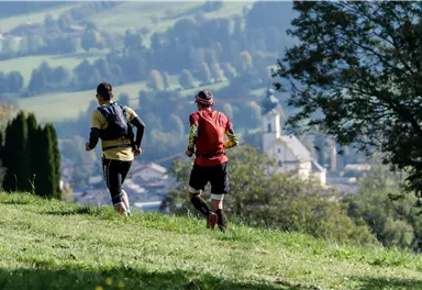 Two runners are jogging across a green meadow. In the background, gentle hills and a small village with a church can be seen.