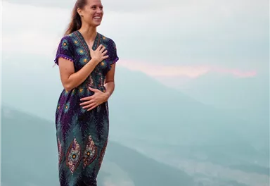 A smiling woman is standing barefoot on a rock in nature. She is wearing a colorful, patterned outfit and looking into the distance.