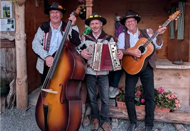 Drei Musiker stehen zusammen und spielen auf ihren Instrumenten. Die Atmosphäre ist freundlich und festlich, mit Blumen im Hintergrund.