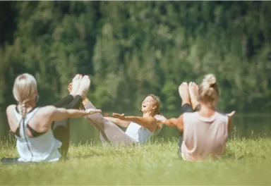 Eine Gruppe von Frauen macht Yoga im Freien. Sie sitzen auf einer Wiese mit Blick auf einen Wald und einen ruhigen See.