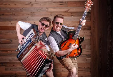 Zwei Musiker posieren fröhlich vor einer Holzwand. Einer spielt das Akkordeon und der andere hält eine Gitarre.