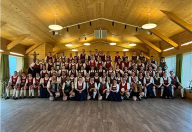 A large group of people in traditional costume stands in a wooden room. They smile and pose together for a photo.