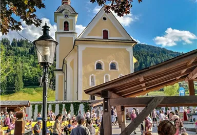 A lively marketplace with many people and a beautiful yellow church in the background. The surroundings are framed by green hills and a blue sky.