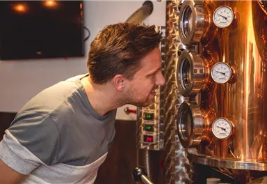 Ein Mann steht vor einer glänzenden Kupferdestille und schaut aufmerksam auf die Messgeräte. Im Hintergrund ist ein Fernseher und industrielles Equipment zu sehen.