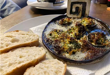 A plate with baked snails and a crispy crust, served on a black plate. Fresh slices of bread are placed alongside.