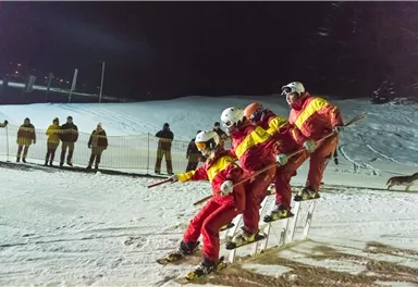 Three skiers in red and yellow suits are making a joint descent in the snow. In the background, spectators can be seen watching the action.
