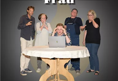Eine Gruppe von fünf Personen reagiert spontan auf eine Szene am Tisch. Eine Person sieht besorgt auf einen Laptop, während die anderen unterschiedliche Emotionen zeigen.