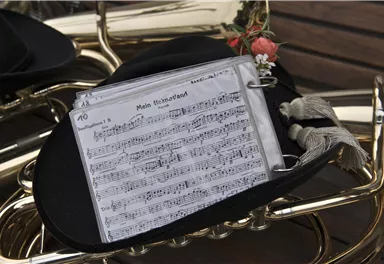 Ein schwarzer Hut mit Notenblättern liegt auf einem musikalischen Instrument. Die Noten zeigen ein Stück namens "Mein Heimatland".