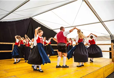 Eine Gruppe von Tänzern in traditionellen Trachten führt einen Volkstanz auf. Sie bewegen sich fröhlich auf einer Bühne in einem Zelt.