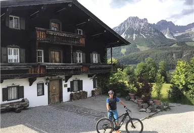 Ein Mountainbiker steht vor einem traditionellen Holzhaus in den Bergen. Im Hintergrund sind die Gipfel und die grüne Landschaft zu sehen.