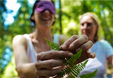 Zwei Frauen in einem Wald, eine trägt eine Augenbinde und hält einen Farn in der Hand. Die andere sieht entspannt zu.