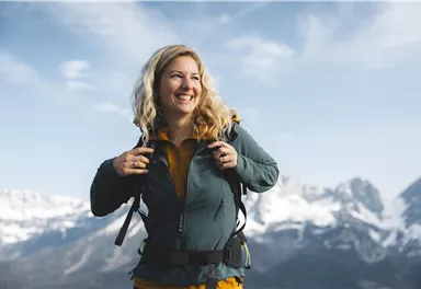 Eine lächelnde Frau mit einem Rucksack steht in den Bergen. Im Hintergrund sehen wir schneebedeckte Gipfel und einen klaren Himmel.