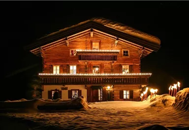 Ein schönes Holzhaus im Schnee, das festlich mit Lichtern dekoriert ist. Fackeln beleuchten den Weg zum Eingang und schaffen eine warme Atmosphäre in der Nacht.