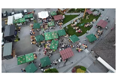 A bustling market with many stalls and green pavilions. Many people are sitting together and enjoying the atmosphere.