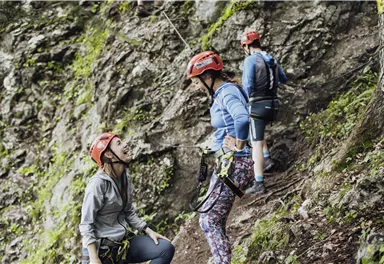 Drei Personen in Kletterausrüstung stehen an einer Felswand. Sie tragen Helme und scheinen eine Kletterroute zu besprechen.