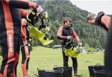 Eine Gruppe von Personen in Neoprenanzügen bereitet sich auf eine Outdoor-Aktivität vor. Einige Teilnehmer organisieren ihre Ausrüstung auf einer Wiese inmitten der Natur.