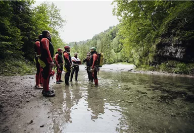 Eine Gruppe von Personen in Neoprenanzügen steht am Ufer eines klaren Gewässers. Die Umgebung ist grün und bewaldet, mit sanften Hügeln im Hintergrund.