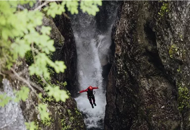 Ein Abenteurer springt von einer Felswand in einen Wasserfall. Umgeben von steilen Felsen und üppigem Grün.