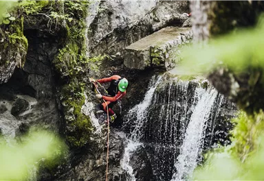 Ein Kletterer in einem grünen Wald klettert an einem Felsen neben einem Wasserfall. Er trägt einen grünen Helm und nutzt ein Seil zur Sicherung.