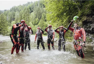 Eine Gruppe von sieben Personen in Neoprenanzügen steht im Wasser und spritzt mit Wasser. Sie haben viel Spaß in einer natürlichen Umgebung mit Bäumen im Hintergrund.
