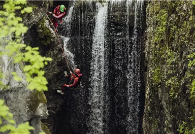 Zwei Personen klettern an einer Felswand neben einem Wasserfall. Die Umgebung ist grün und naturnah.