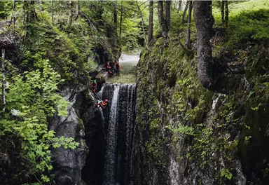 Ein beeindruckender Wasserfall, umgeben von grünen Bäumen, fließt durch eine enge Schlucht. Einige Personen sind in der Nähe des Wassers aktiv.