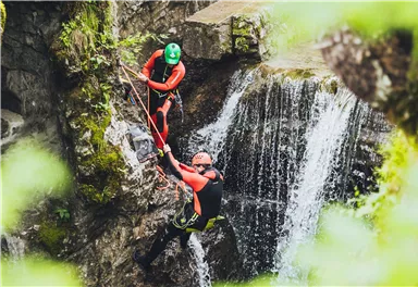 Zwei Personen in Neoprenanzügen, die an einer Felswand klettern. Im Hintergrund ist ein Wasserfall zu sehen.