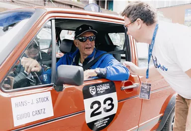 An older man is sitting in a red car, wearing sunglasses and a cap. Another man is standing outside next to the car and talking to him.