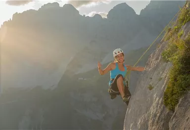 A person is climbing a steep rock face in the mountains. The scene is illuminated by sunlight and showcases the joy of climbing.