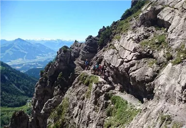Eine Gruppe von Wanderern geht auf einem schmalen Pfad entlang steiler Felsen. Im Hintergrund erstreckt sich eine atemberaubende Berglandschaft.