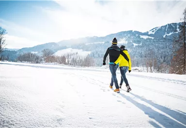 Ein Paar geht durch eine verschneite Landschaft. Die Sonne scheint und die Berge sind im Hintergrund sichtbar.