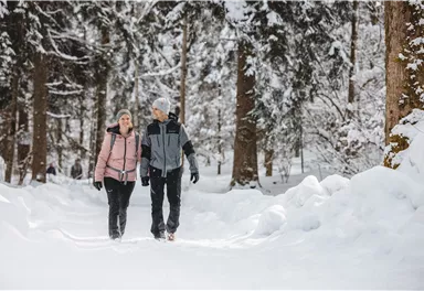 Ein Paar geht lächelnd durch eine verschneite Waldlandschaft. Die Bäume sind mit Schnee bedeckt und der Weg ist gut sichtbar.