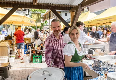 A joyful outdoor event with a market stall. Two people, smiling warmly, stand at the stall and serve food.