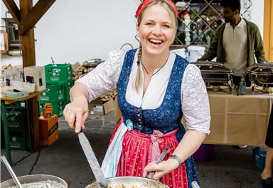 A cheerful woman in traditional attire is cooking at the market. She smiles and serves a delicious dish.