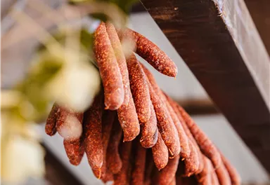 A selection of spicy sausages hangs on a wooden shelf. Blurred objects can be seen in the background.