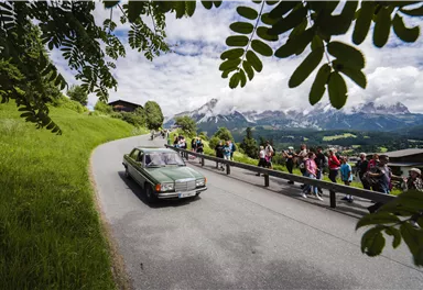 Eine Gruppe von Menschen wandert auf einem Weg durch eine grüne Landschaft. Im Hintergrund sind Hügel und ein kleiner Ort zu sehen.