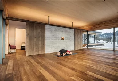 A minimalist room with wooden floors and large windows. In the center, a person is practicing yoga.