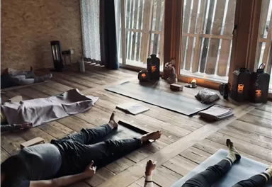 Ein ruhiger Raum mit Holzfußboden, in dem Menschen entspannt auf Yogamatten liegen. Sanftes Licht strömt durch die Fenster und schafft eine entspannende Atmosphäre.