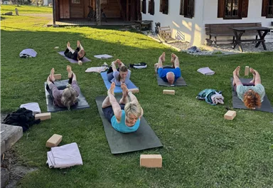 Eine Gruppe von Menschen macht Yoga draußen auf einer Wiese. Im Hintergrund sieht man ein traditionelles Holzhaus und einen klaren Himmel.