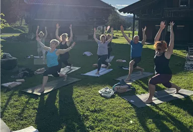 Eine Gruppe von Menschen macht Yoga im Freien unter einem klaren Himmel. Die Sonne scheint, und die Atmosphäre ist entspannt.
