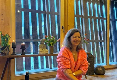 Eine Frau sitzt im Lotussitz auf einer Matte in einem gemütlichen Raum. Im Hintergrund sind Pflanzen und große Fenster mit Blick auf die Natur zu sehen.
