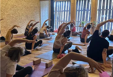 Eine Gruppe von Menschen praktiziert Yoga in einem hellen Raum mit Holzwänden. Sie dehnen sich auf Matten und nutzen Yogablöcke.