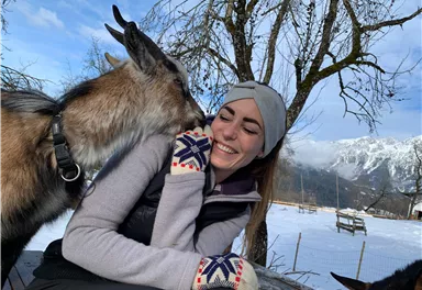 A woman smiles happily as a goat kisses her. In the background, snow-covered mountains and trees can be seen.