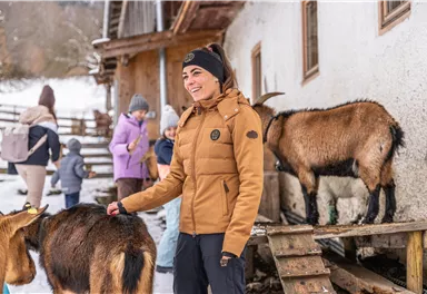 Eine Person mit brauner Jacke steht vor einem Stall und streichelt eine Ziege. Im Hintergrund sind weitere Personen zu sehen, die sich ebenfalls mit Ziegen beschäftigen.