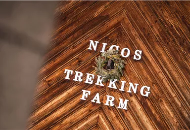 Ein Holzboden mit dem Schriftzug "Nicos Trekking Farm" und einem grünen Kranz. Die Atmosphäre ist rustikal und einladend.