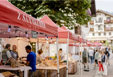 Ein lebendiger Marktplatz bei Dämmerung mit Ständen, die lokale Produkte anbieten. Menschen schlendern durch die Gassen und genießen die Atmosphäre.