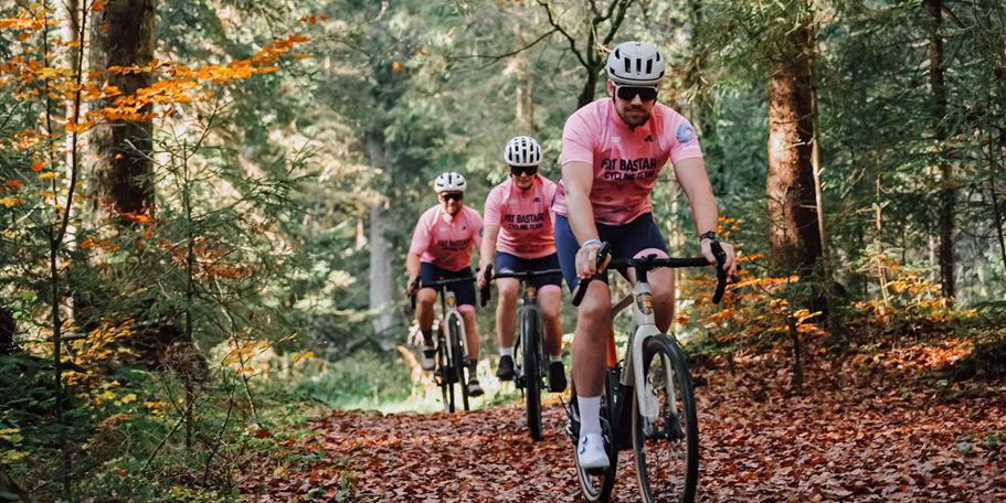 Group of cyclists in pink jerseys riding through a forest path. The ground is covered with autumn leaves, while the trees in the background glow.