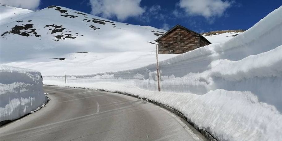 A snow-covered road surrounded by high snow banks. In the background stands a small wooden house under a blue sky with some clouds.