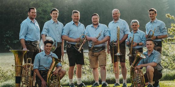 Eine Gruppe von Musikern steht im Freien und trägt traditionelle Trachten. Sie halten verschiedene Blasinstrumente und lächeln in die Kamera.